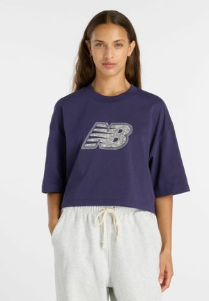 Frau mit langen braunen Haaren, die ein marineblaues, kurzes T-Shirt mit weißem und silbernem "NB"-Logo und hellgraue Jogginghose mit Kordelzug trägt.