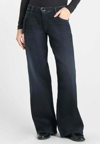 Jeans bleu foncé à jambes larges avec une finition en denim texturé, présentant une poche avant et des embellissements en métal argenté à la taille.