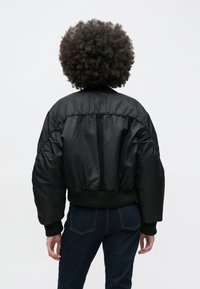 Persoană cu păr creț purtând o jachetă neagră puf și blugi închiși la culoare, stând cu spatele la un fundal alb simplu.