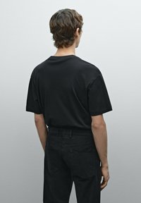 Sort bomulds T-shirt med afslappet pasform, korte ærmer og rund halsudskæring, sammen med sorte bukser, set bagfra.