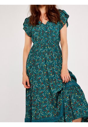 Femme portant une robe bleu sarcelle à manches courtes et longueur genou, avec un motif floral en rouge, jaune et blanc, tenant une partie de la jupe.