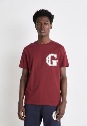Camiseta de algodón burdeos con cuello redondo, mangas cortas y un gran logo en blanco "GANT G" en el pecho. Ajuste simple y casual.