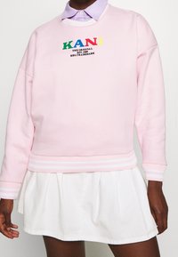 Sudadera rosa con el texto "KANI" en letras multicolores. Presenta puños y dobladillo acanalados con una falda blanca. Material de algodón suave.