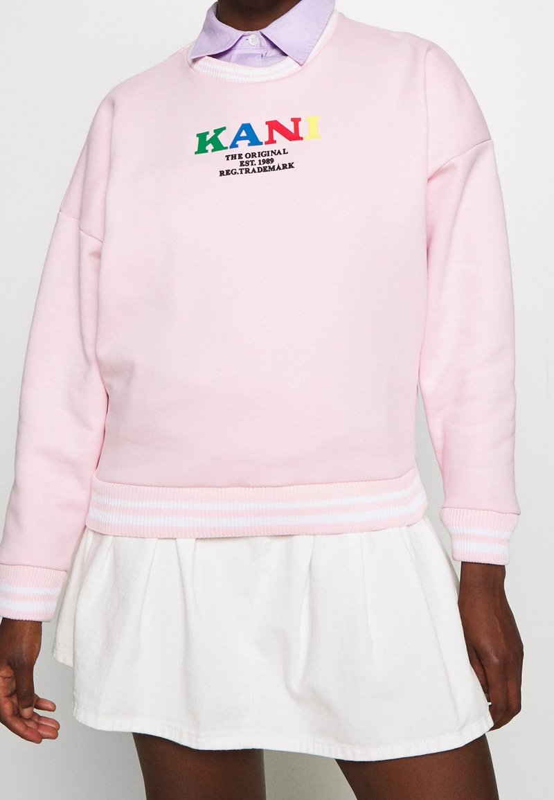 Sudadera rosa con el texto "KANI" en letras multicolores. Presenta puños y dobladillo acanalados con una falda blanca. Material de algodón suave.