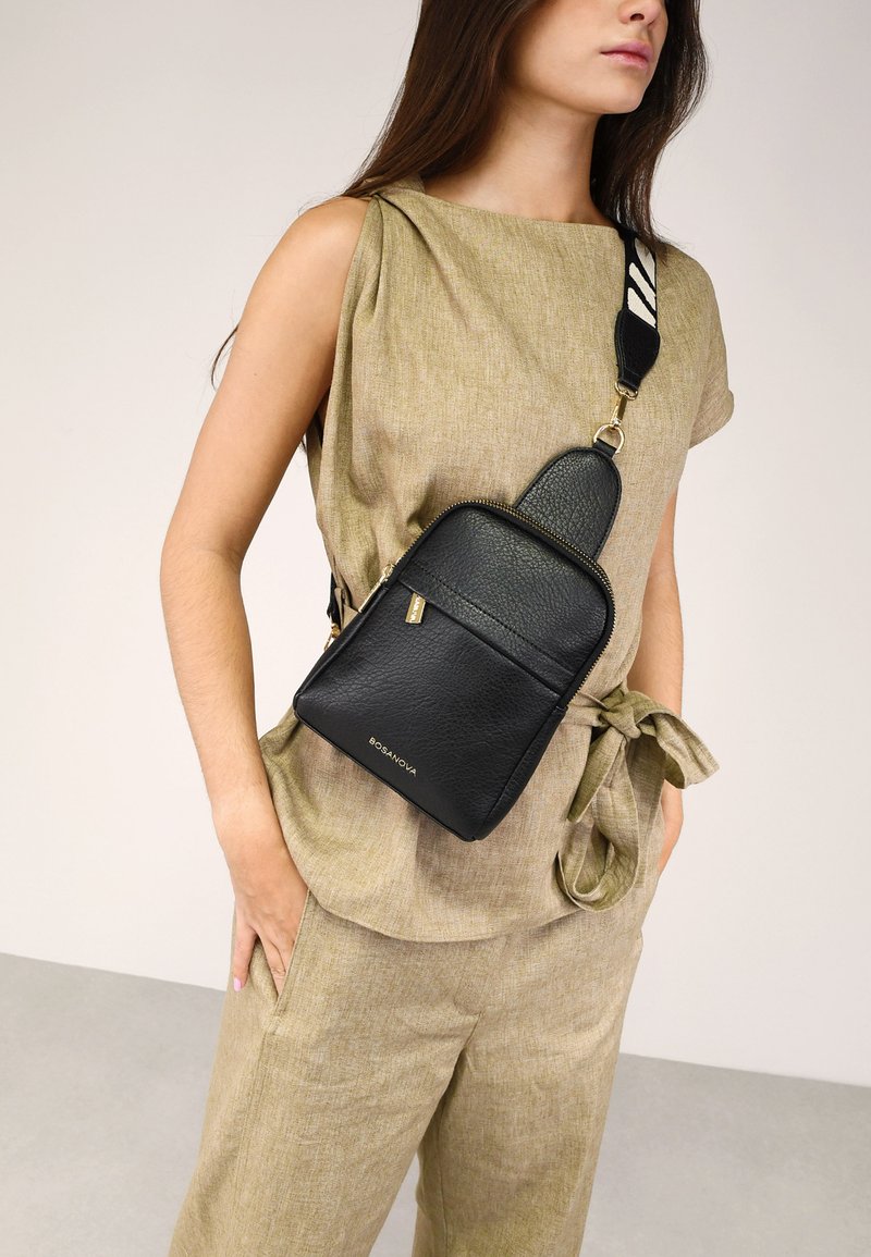 Bolso cruzado de cuero negro con una superficie texturizada, cierre de cremallera y una correa desmontable con un acento blanco de patrón.