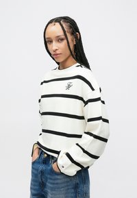 Weißes Langarmoberteil mit kräftigen schwarzen Streifen, versehen mit einem kleinen, gestickten Logo auf der Brust und Bündchen mit kontrastfarbigen Akzenten.