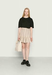 Jovem mulher vestindo uma blusa preta de manga curta, saia bege listrada com babados, sapatos pretos, segurando uma bolsa tote bege, de pé em um chão cinza com uma parede clara.