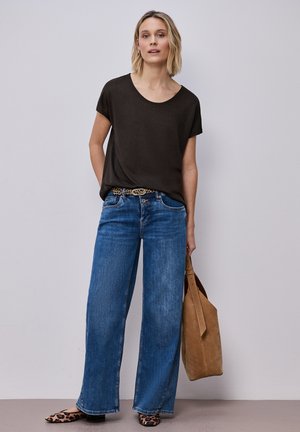 Donna bionda che indossa una maglietta nera a maniche corte, jeans blu a gamba larga con cintura leopardata, ballerine leopardate, che tiene in mano una borsa tote in camoscio marrone.