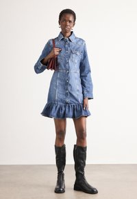 Vestido camiseiro de ganga com botões à frente, mangas longas e bainha franzida. Usado com botas pretas até ao joelho e uma pequena mala vermelha.