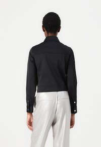 Veste noire courte à manches longues, détail sur le yoke avant, poignets boutonnés, portée avec un pantalon large argenté brillant. Texture de tissu lisse.