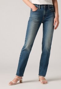 Blauwe denim rechte broek met een vervaagd uiterlijk, vijf zakken en een knoopsluiting. Gecombineerd met witte bandjesandalen.