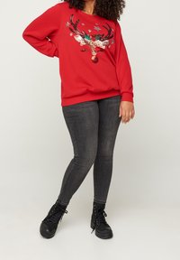 Rode sweater met een pailletten rendierdesign en kleurrijke sterren aan de voorkant, gecombineerd met donkere strakke jeans en zwarte veterlaarzen.