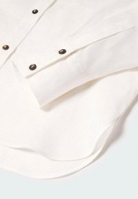 Hvid langærmet skjorte i manchet med mørkebrune knapper, foldet på en lys overflade, viser knapdetaljer og tekstur i stoffet.