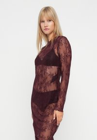 Jauna sieviete ar taisnu blondiem matiem, ģērbusies caurspīdīgā daļēji sarkanas mežģīņu kleitā pār atbilstošiem krūštura un apakšveļas komplektiem, stāv uz balta fona.