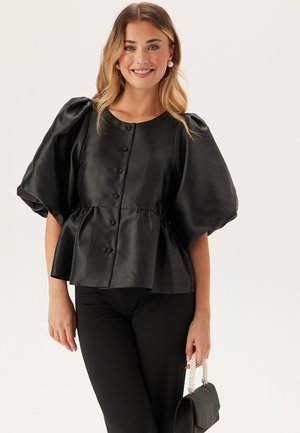Černá saténová blůza s nafouklými rukávy, kulatým výstřihem a předním zapínáním na knoflíky. Peplum styl s texturovaným povrchem, kombinována s černými kalhotami.