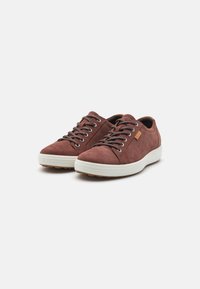 Une paire de baskets basses en daim brun avec des lacets gris et des semelles en caoutchouc blanc sur un fond blanc.