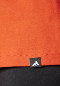 Narančasta pamučna majica s ravnim rubom i crnom adidas oznakom na donjem lijevom kutu, s finom rebrastom teksturom.