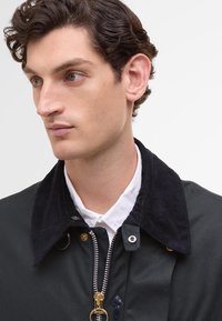 Casaco preto com colarinho de veludo cotelê, fecho éclair prateado e detalhes em metal dourado. Camisa branca visível por baixo. Textura de tecido suave.