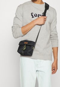 Persoon in een lichtgrijze sweater met de tekst "forêt" en lichtblauwe jeans, die een zwarte MSGM schoudertas met groene karabijnhaak draagt.