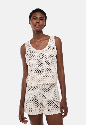Femme portant un haut sans manches en crochet couleur crème et un short assorti sur un fond clair uni.