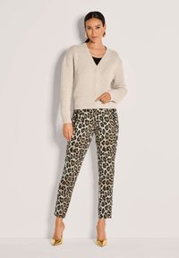 Frau in beiger Strickjacke, schwarzem Top, Leopardenmuster-Hose, goldenen Absätzen und goldenen geometrischen Ohrringen steht vor einer hellen Wand.