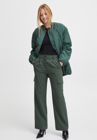 Zelená bomber bunda s hladkou, lesklou texturou; černý přiléhavý top; zelené široké cargo kalhoty s bočními kapsami; černé lodičky.