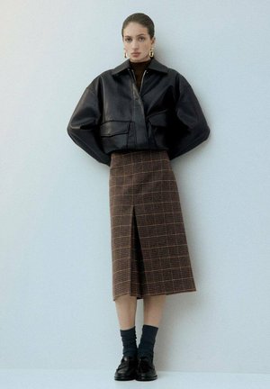 Veste courte en cuir noir avec de grandes poches, associée à une jupe en tartan marron arrivant au genou avec une fente sur le devant. Des chaussettes et des chaussures sombres complètent le look.