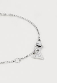 Bracciale in metallo argentato con una charm triangolare incisa con "GUESS?" e chiusura a moschettone. Texture liscia.