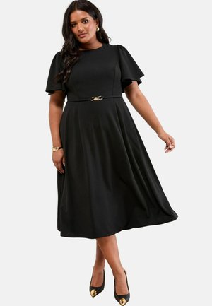 Schwarzes Midi-Kleid mit ausgestellten kurzen Ärmeln, einer Taillenbetonung mit goldenen Details und einer glatten Textur. Kombiniert mit schwarzen High Heels mit goldenen Spitzen.