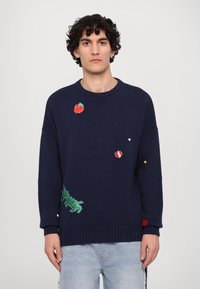 Marineblauer Pullover mit gestickten Motiven, darunter eine Erdbeere, ein roter Ball und grüne Blätter. Grobstrick-Textur mit gerippten Bündchen.