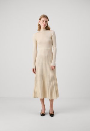 Strickkleid in hellem Beige mit einem enganliegenden Langarm-Oberteil und einem gefältelten Rock. Verfügt über Schulter-Ausschnitte und eine glatte Textur, kombiniert mit schwarzen Absätzen.