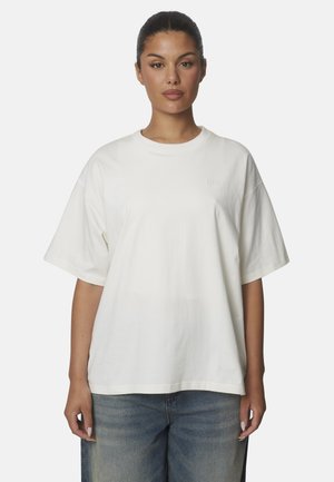 Vrouw met donker haar naar achteren gebonden, die een eenvoudige witte oversized korte mouw T-shirt en blauwe spijkerbroek draagt, staand tegen een lichte achtergrond.