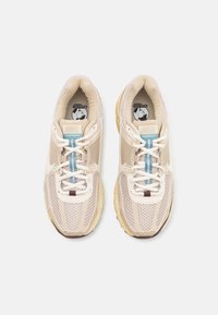Baskets de sport beiges avec une tige en mesh, accents bleus et semelle beige. Dotées d'un laçage et d'un branding subtil sur la semelle intérieure.