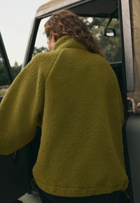 Maslinasto zelena flis jakna s visokim ovratnikom, oversized kroja, teksturiranom tkaninom i vezicom na rubu, koju nosi osoba koja ulazi u vozilo.