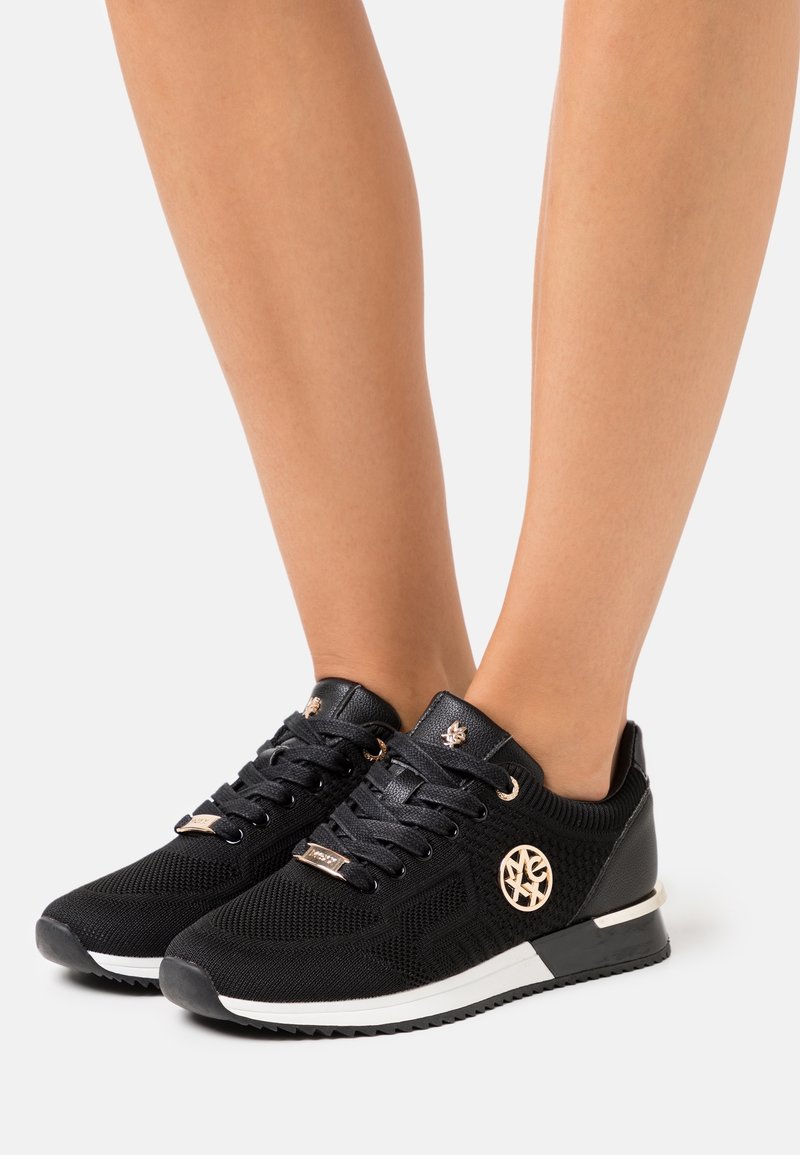 Baskets noires avec une tige en maille, des accents texturés et un emblème métallique sur le côté ; dotées de lacets noirs et d'une semelle extérieure blanche.