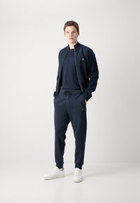 Námornícky športový komplet s bundou s zipsom a tapered nohavicami, vyrobený z hladkej tkaniny, s elastickými manžetami a sťahovacím pásom. Biele tenisky.
