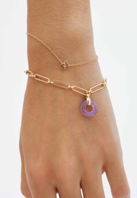 Pulsera de cadena de oro con eslabones alargados, un charm circular morado y un tobillo delgado con un acento en forma de corazón. Piel texturizada visible.