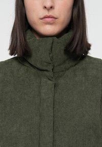 Vrouw met steil bruin haar, gekleed in een olijfgroene ribgebreide trui met hoge hals, crop tot onder de neus, neutrale uitdrukking.