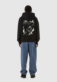Svart hettegenser med en grafisk design som har en krone og et ansikt på ryggen. Kombinert med løstsittende blå denimjeans og svarte sko.