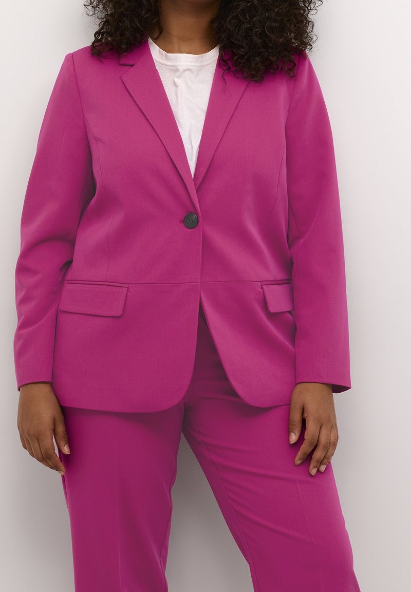 Mujer con cabello rizado vestida con una chaqueta de traje rosa brillante y pantalones a juego sobre una camisa blanca, frente a un fondo neutro.