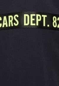 Navy fabric with a matte texture features a black woven band labeled "CARS DEPT. 82" in bright yellow lettering.