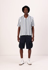 Camisa de manga corta a rayas en azul y blanco, combinada con pantalones cortos de carga en azul marino con bolsillos laterales, usados con zapatillas blancas y un sombrero negro.