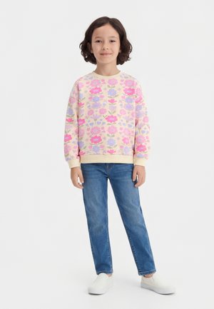 Fille debout portant un sweat-shirt crème avec des fleurs roses et violettes, un jean bleu et des chaussures blanches sans lacets sur fond blanc.