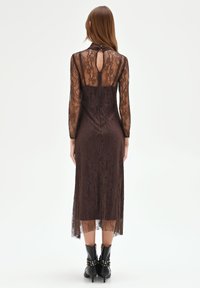 Bruine kanten jurk met lange mouwen, transparante achterkant en een sleutelgatdetail. Lengte net onder de knieën met een getailleerd silhouet en een textuurpatroon.