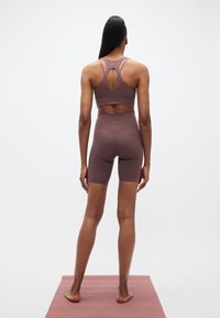 Καφέ αθλητικ ό σουτιέν με λουριά που διασταυρώνονται στην πλάτη και αντίστοιχο σορτς με ψηλή μέση, κατασκευασμένο από λείο, ελαστικό υλικό, παρουσιασμένο με φόντο ουδέτερο.