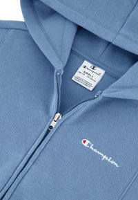 Felpa con zip di colore azzurro chiaro realizzata in un misto di cotone con una texture morbida. Presenta il logo Champion in bianco e rosso sul petto e un'etichetta interna.