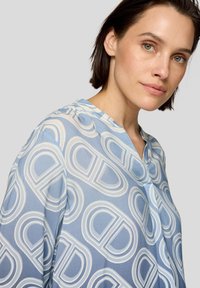 Himmelbluse mit einem sich wiederholenden weißen kreisförmigen Muster, V-Ausschnitt und glatter Textur. Sichtbare Nähte am Kragen.