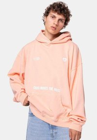 Sudadera rosa claro con un gran bolsillo frontal, texto gráfico "EL CAOS HACE A LA MUSA" y un pequeño logo de corazón en el pecho. Tejido de mezcla de algodón.