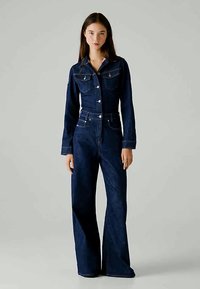 Tume teksajumpsuit pikkade varrukatega, laienevate sääretega, rinna taskutega, metallnuppudega ja kohandatud vöökohaga. Sile tekstuur ja klassikaline disain.