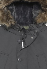 Grijze parka met een capuchon gevoerd met nepbont. Voorzien van een ritssluiting, drukknoopsluiting en twee voorzakken. Materiaal lijkt glad met een gedempte textuur.